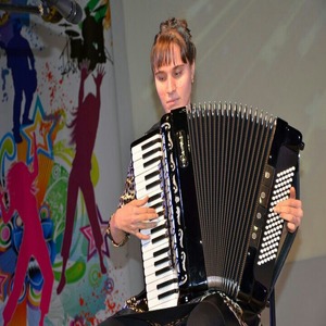 Карина Райская играет на аккордионе, слева от нее расписанная силуэтами людей стена