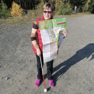 Маргарита Мельникова стоит на дорожке у опушки леса, держа в руках карту с надписью "Майская прогулка"