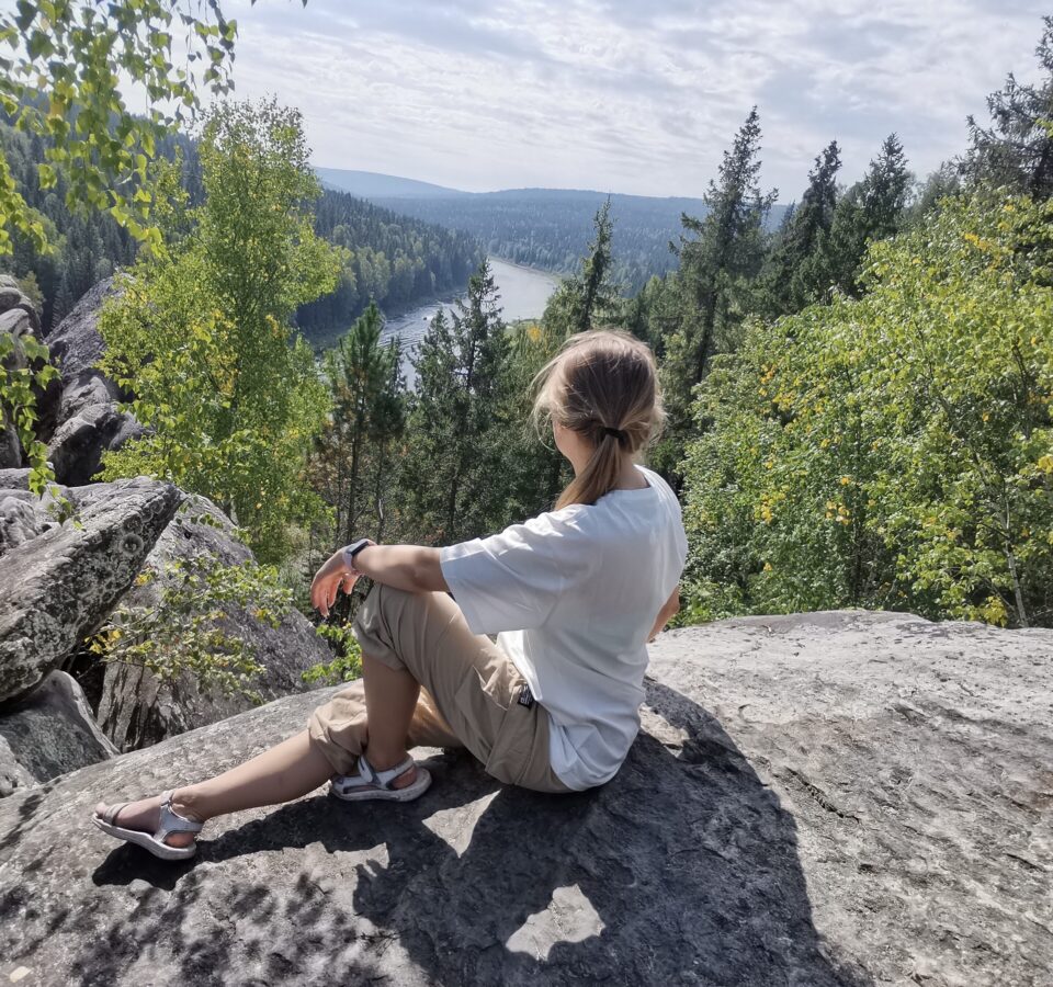 Анна Крапивина сидит на скале спиной к фотографу, смотрит на лес и реку перед ней
