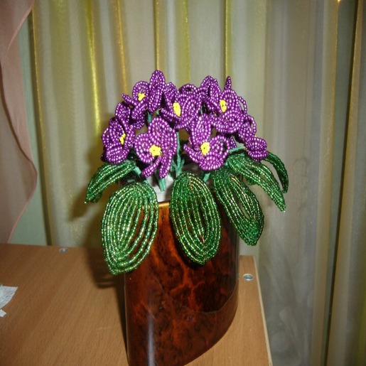 На столе в коричневой вазе стоит букет фиолетовых ярких цветов из бисера
