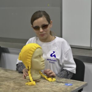 Лариса Малышкина сидит за столом в футболке с символикой "Абилимпикса", перед ней лежит трехмерная модель головы в вязаной желтой шапке