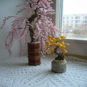 На подоконнике стоит сделанное из нежно-розового бисера деревце, напоминающее сакуру. Рядом другое деревце, декорированное желтоватыми камешками