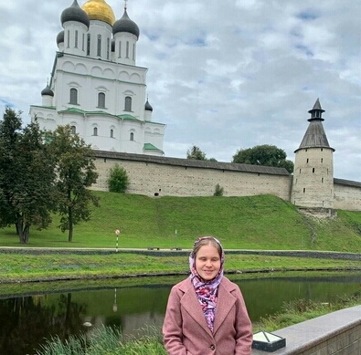 Улыбающаяся Ольга Александрова в бежевом пальто и платке на голове стоит на берегу небольшой речки, сразу за рекой на холме виден белокаменный православный храм.