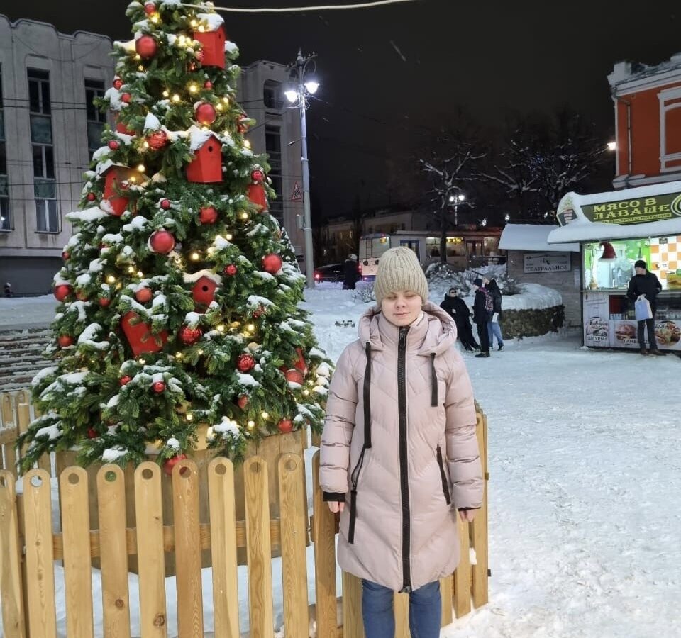 Алиса Любимова стоит на заснеженной площади у наряженной новогодней ёлки