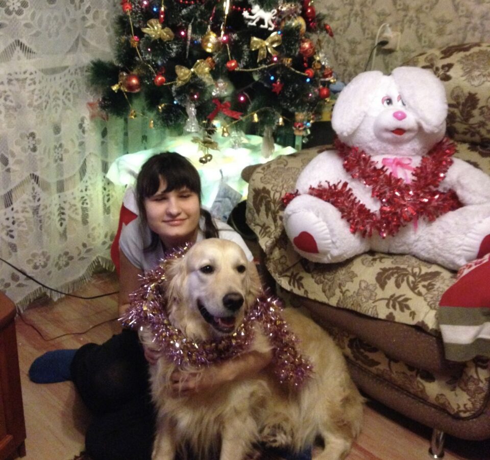 Николь Антонова сидит в комнате на полу под новогодней ёлочкой и обнимает собаку – золотистого ретривера, обмотанного мишурой