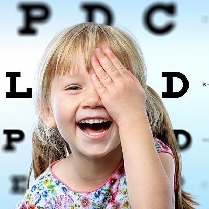 Смеющаяся девочка закрывает одной рукой левый глаз, сзади нее на стене видна таблица для проверки зрения, верхняя и нижняя строка которой слегка размыты