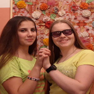 Ольга Александрова с подругой стоят, держась за руки, на фоне стены с цветами