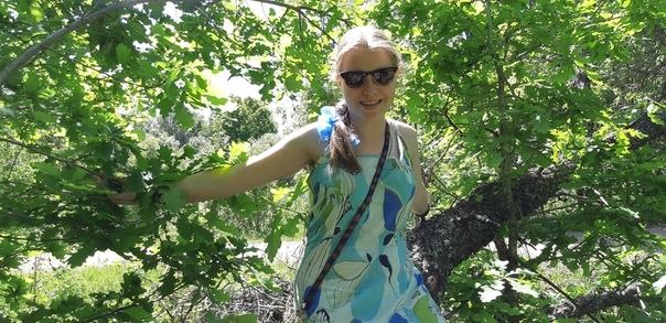 Ольга Александрова в легком платье сидит на стволе дерева на фоне листвы