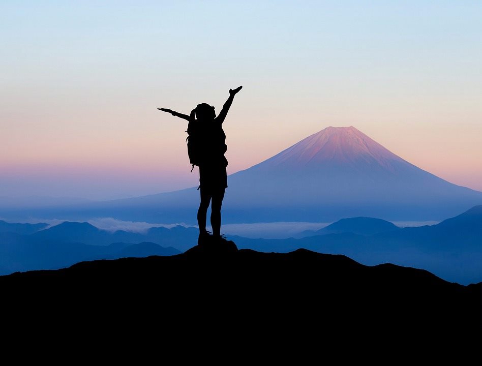 На фоне заката в горах человек с рюкзаком, подняв руки, радуется восхождению на вершину