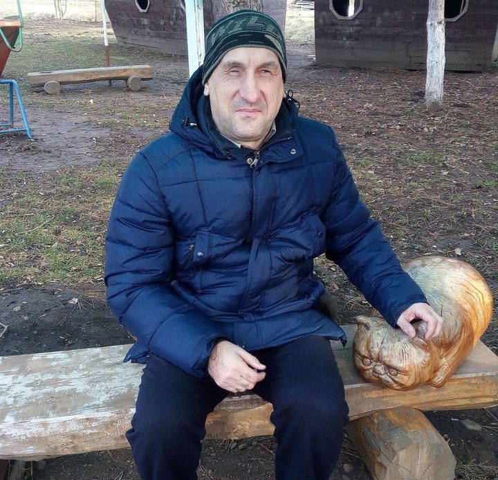 Олег сидит на скамейке возле вырезанного из дерева декоративного кота