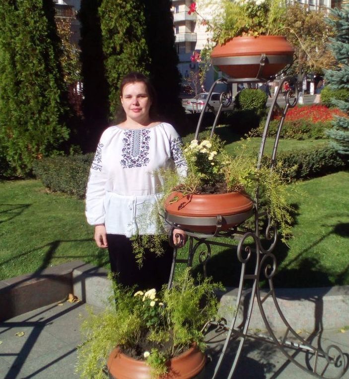 Татьяна Комарницкая стоит возле декоративной подставки с вьющимися растениями в парке