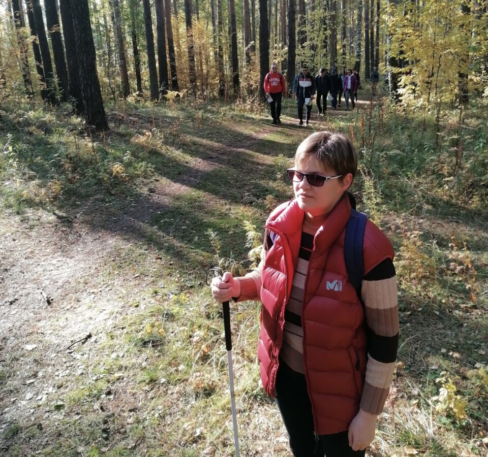 Маргарита Мельникова с рюкзаком за спиной стоит в лесу на краю тропы, позади нее группа людей, идущих к ней