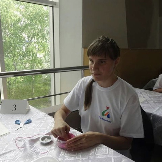 Карина сидит за столом в футболке с символикой "Абилимпикса", на столе перед ней баночка с бисером