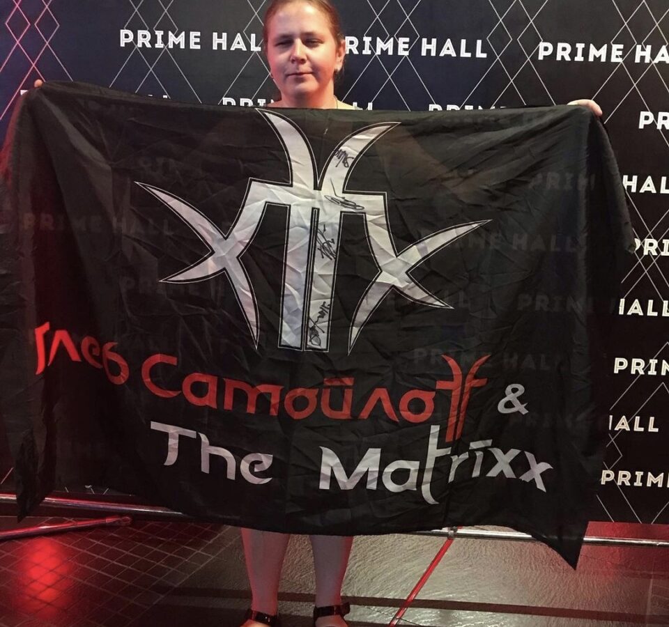 Инна Новик показывает огромный черный флаг, закрывающий всю ее фигуру, на флаге надпись: "Глеб Самойлов и The Matrixx"
