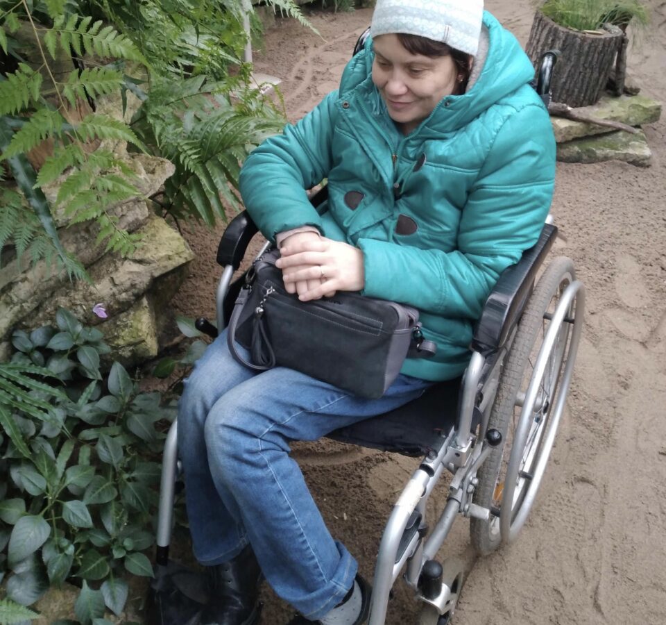 Наталья Демьяненко в инвалидной коляске рядом с высоким растением в оранжерее ботанического сада