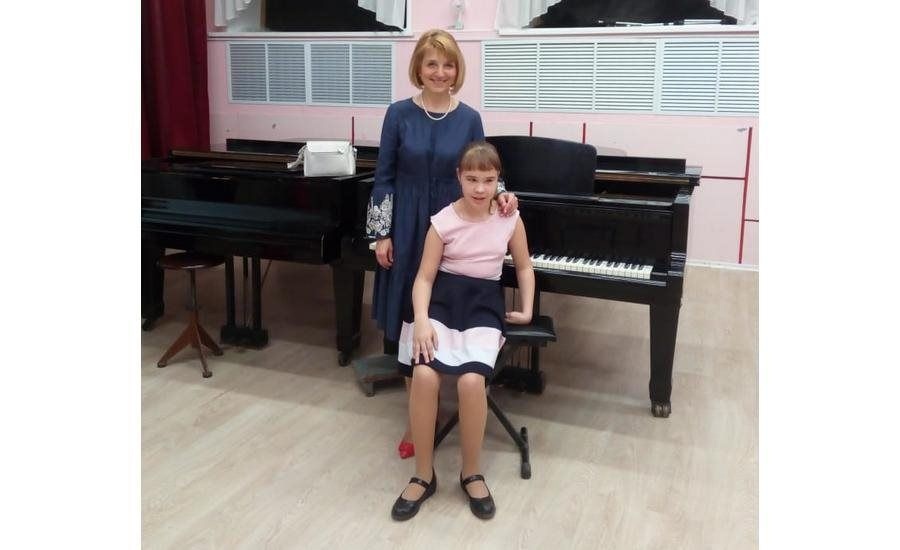 Надежда Иванова сидит на стульчике спиной к фортепиано, сзади нее стоит учительница, положив руку ей на плечо, и улыбается
