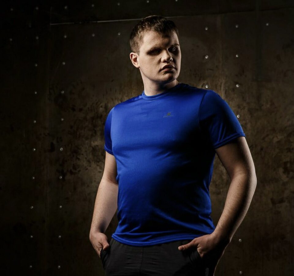 Алексей Лапшинов в синей футболке на фоне тёмной стены