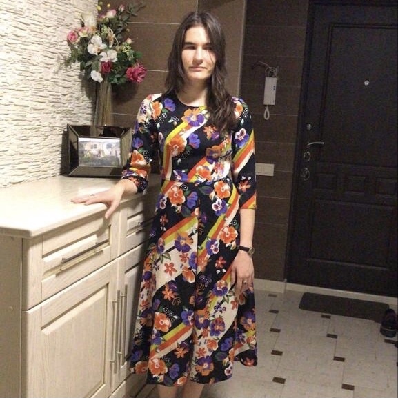 Валерия Артемова в разноцветном платье стоит в прихожей квартиры, позади нее на тумбочке букет цветов